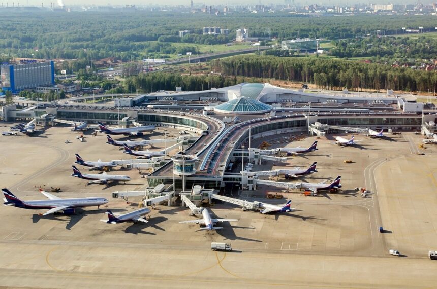 Vista aérea de un gran aeropuerto con numerosos aviones de Aeroflot estacionados en plataforma junto a la terminal, con la ciudad y bosques de fondo