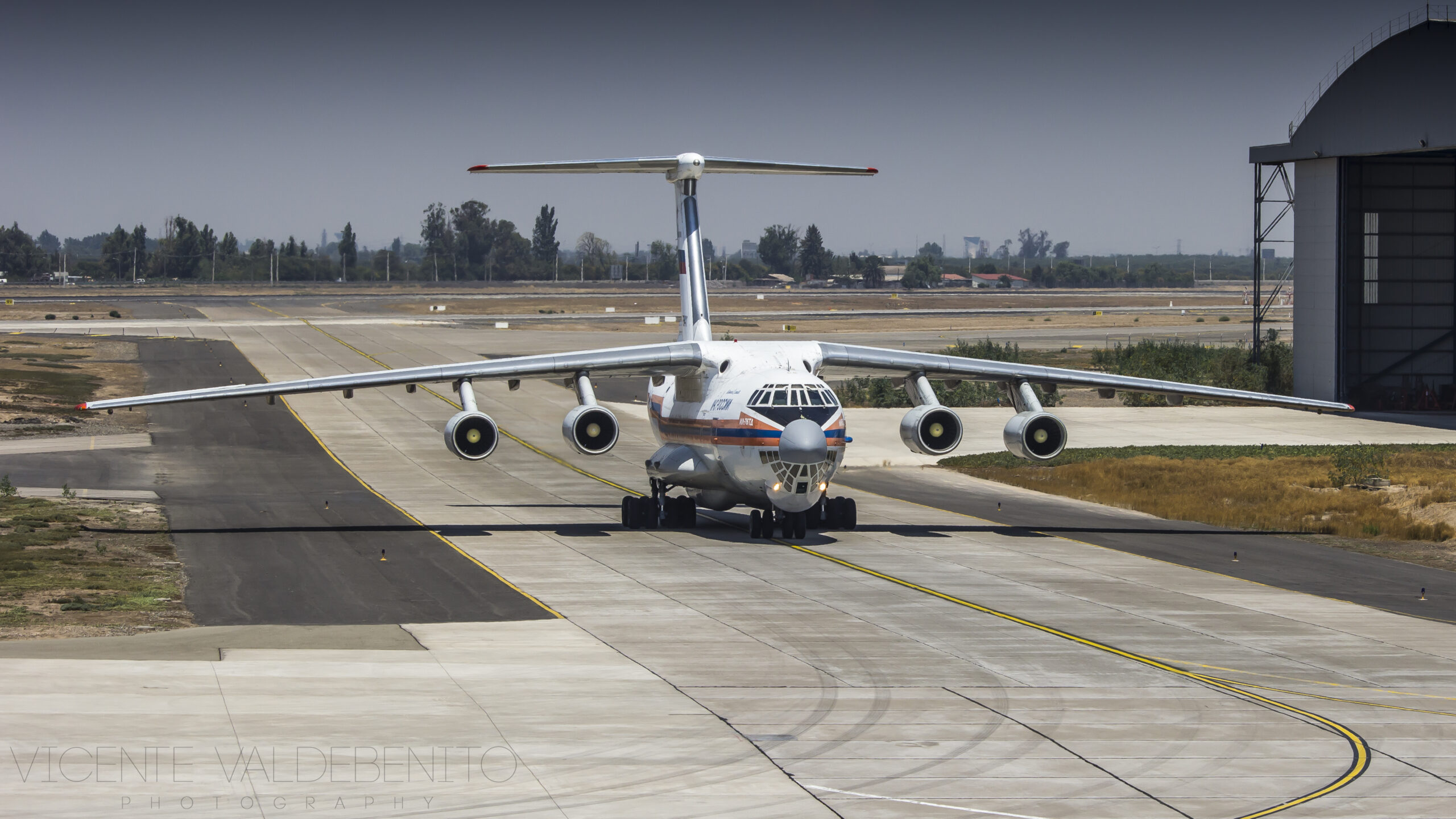 vión cuatrimotor Ilyushin Il-76 de fabricación rusa, con librea blanca, azul y naranja y marcas rusas, rodando en plataforma junto a un hangar