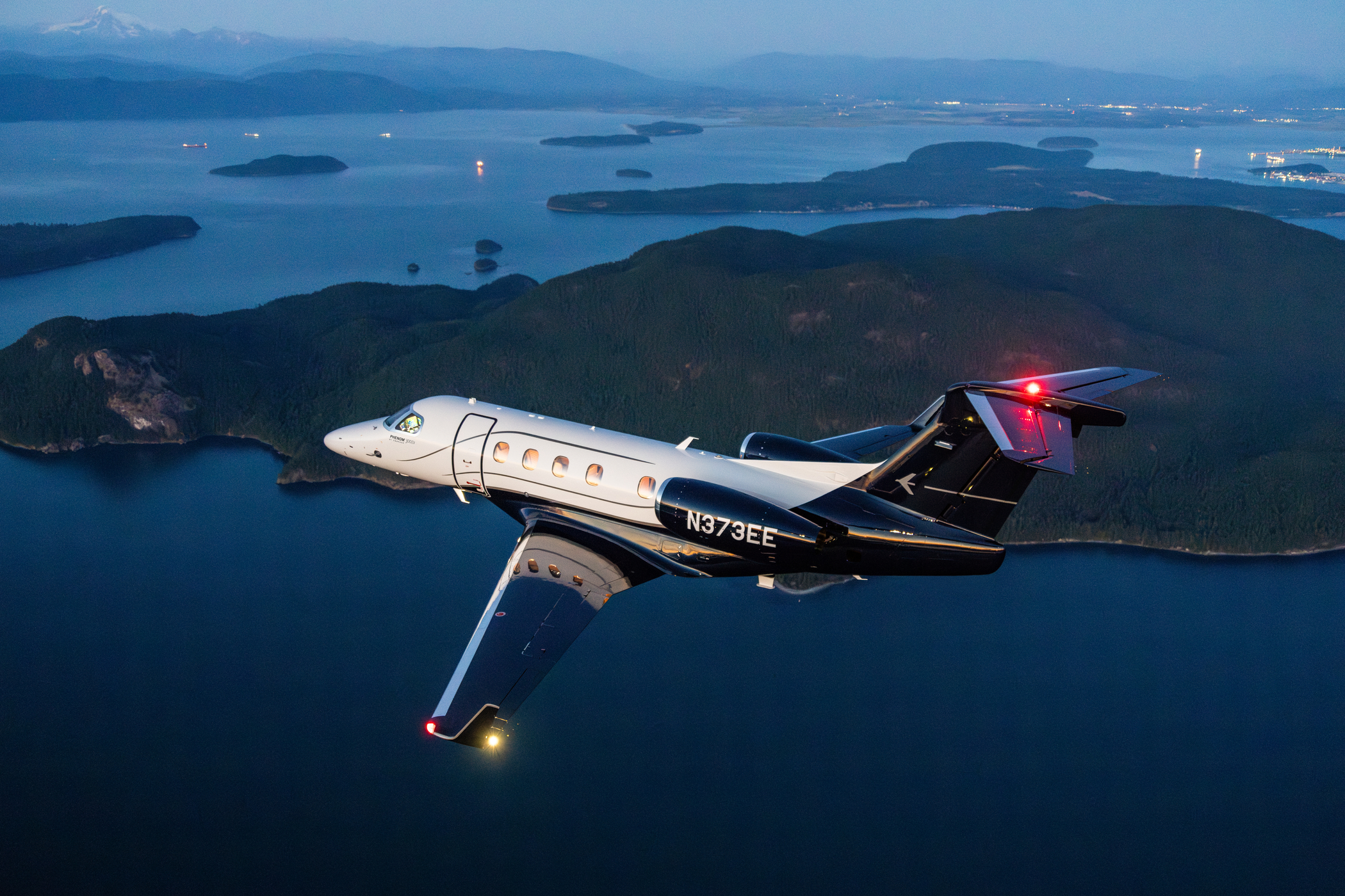 Jet ejecutivo Embraer Phenom 300 matrícula N373EE en vuelo al atardecer, con librea blanca y negra, sobre un paisaje de islas y agua