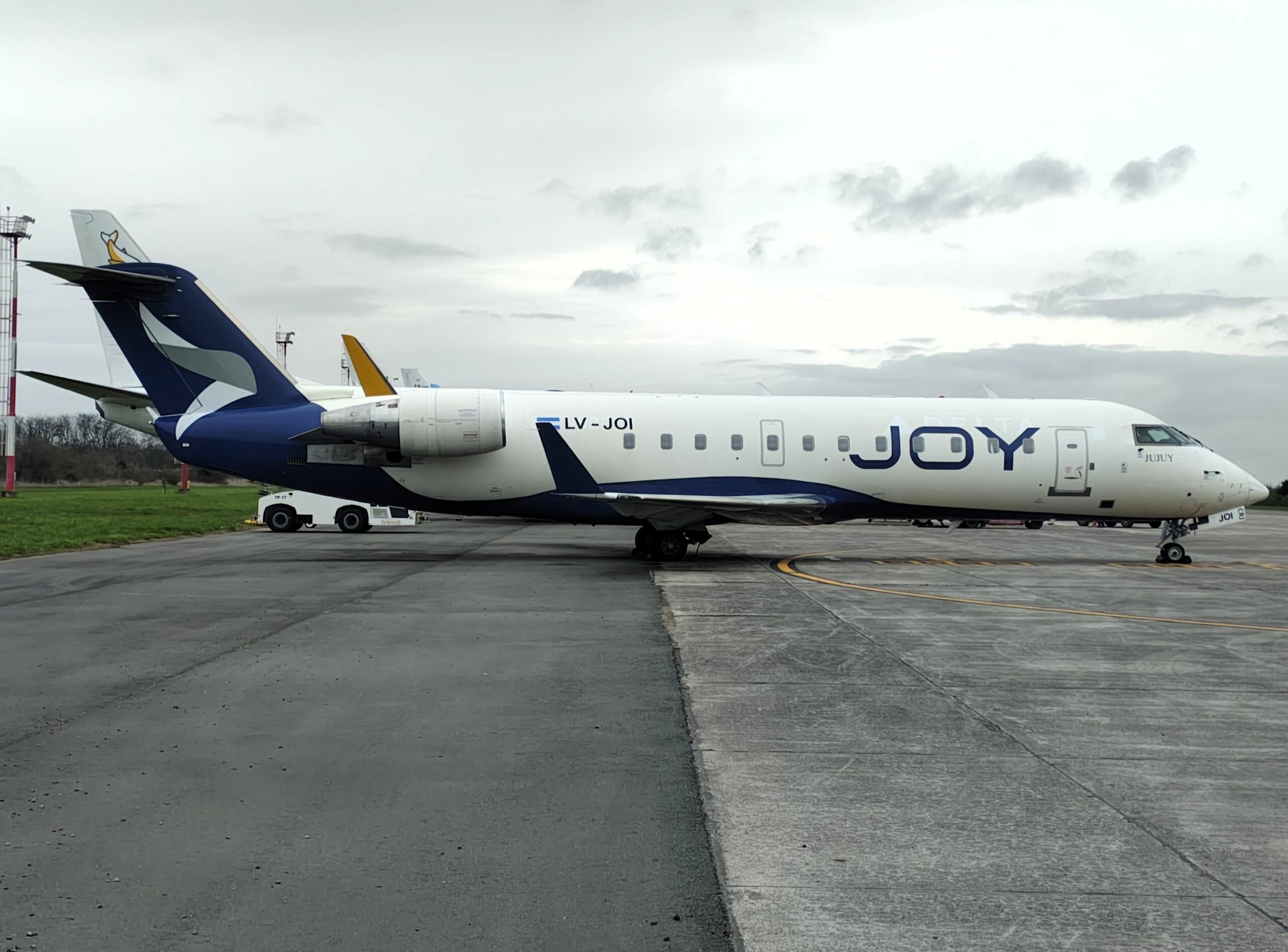 Bombardier CRJ200 LV-JOI de Joy Airline con librea azul y blanca estacionado en plataforma, con titulado JOY en el fuselaje y un tractor de remolque en el tren de nariz