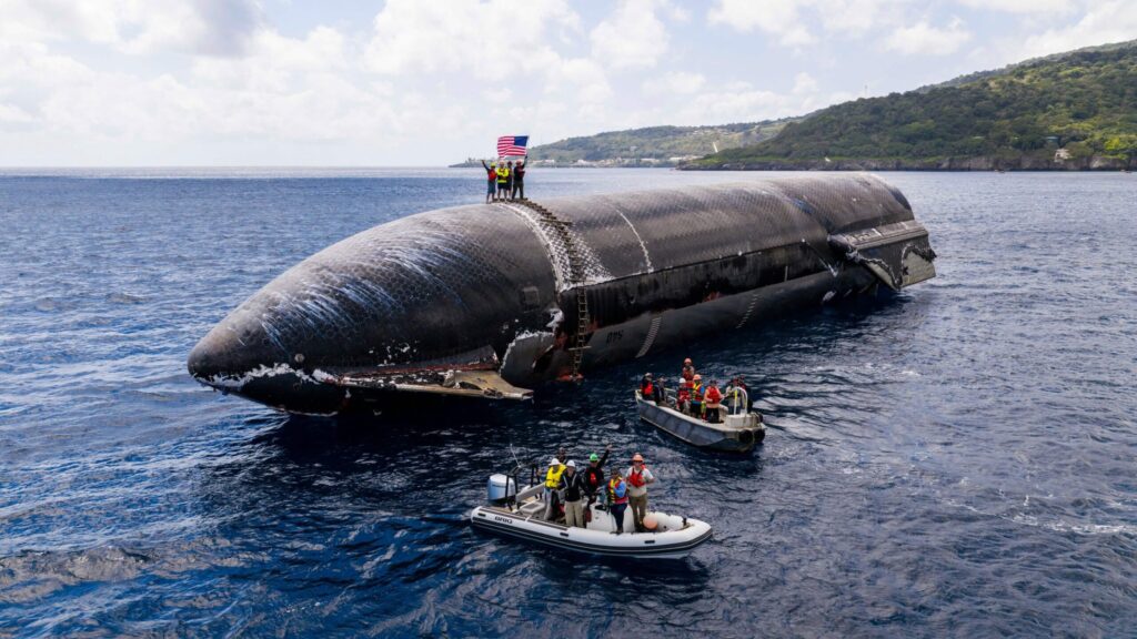 Etapa superior de una Starship flotando en el océano con el escudo térmico de losetas negras a la vista, personas de pie sobre ella con una bandera y botes de recuperación alrededor, con una isla de fondo
