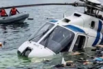 Helicóptero blanco con franjas azules parcialmente sumergido en el lago de Atitlán, Guatemala, con restos flotando alrededor y rescatistas con chalecos naranja en una embarcación inflable al fondo