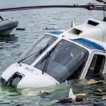 Helicóptero blanco con franjas azules parcialmente sumergido en el lago de Atitlán, Guatemala, con restos flotando alrededor y rescatistas con chalecos naranja en una embarcación inflable al fondo