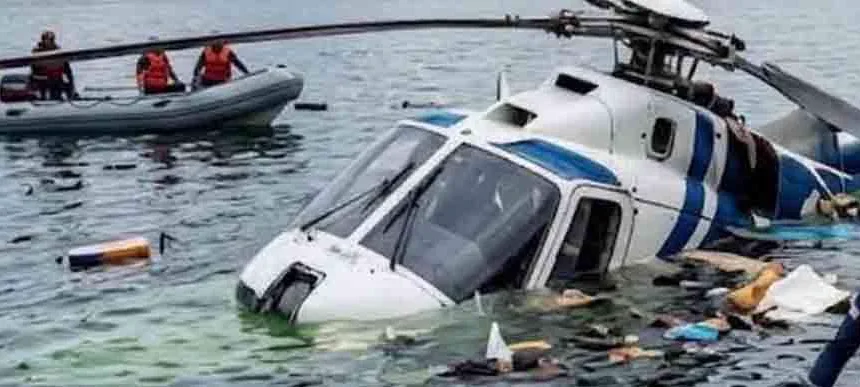 Helicóptero blanco con franjas azules parcialmente sumergido en el lago de Atitlán, Guatemala, con restos flotando alrededor y rescatistas con chalecos naranja en una embarcación inflable al fondo